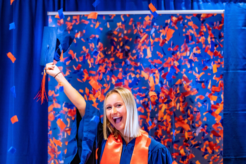 Graduate in regalia joyfully holding up a graduation cap while standing under a shower of orange and blue confetti.