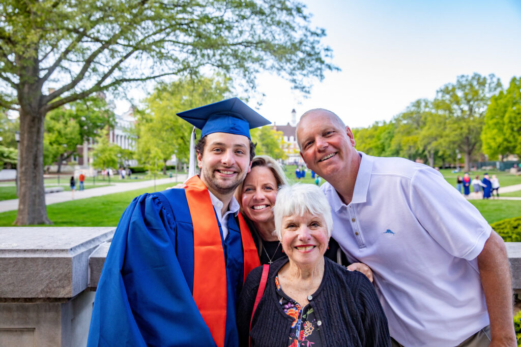 A graduate in orange and blue regalia joyfully poses with three family members on a university of Illinois main quad.