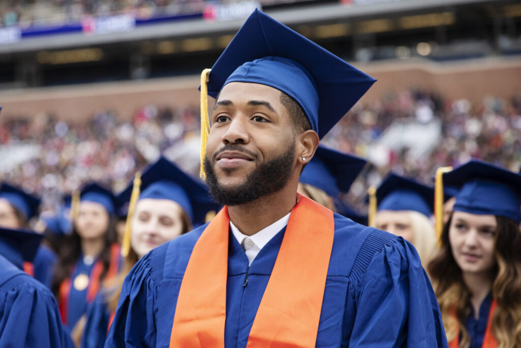 Graduating student in orange and blue regalia smiling at a university commencement ceremony, surrounded by fellow graduates.