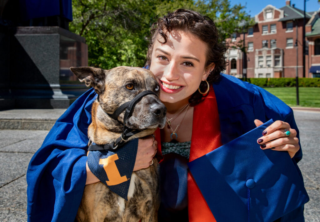 A person in regalia holding a graduation cap and smiling while embracing a dog wearing a bandana with a Block I on campus.