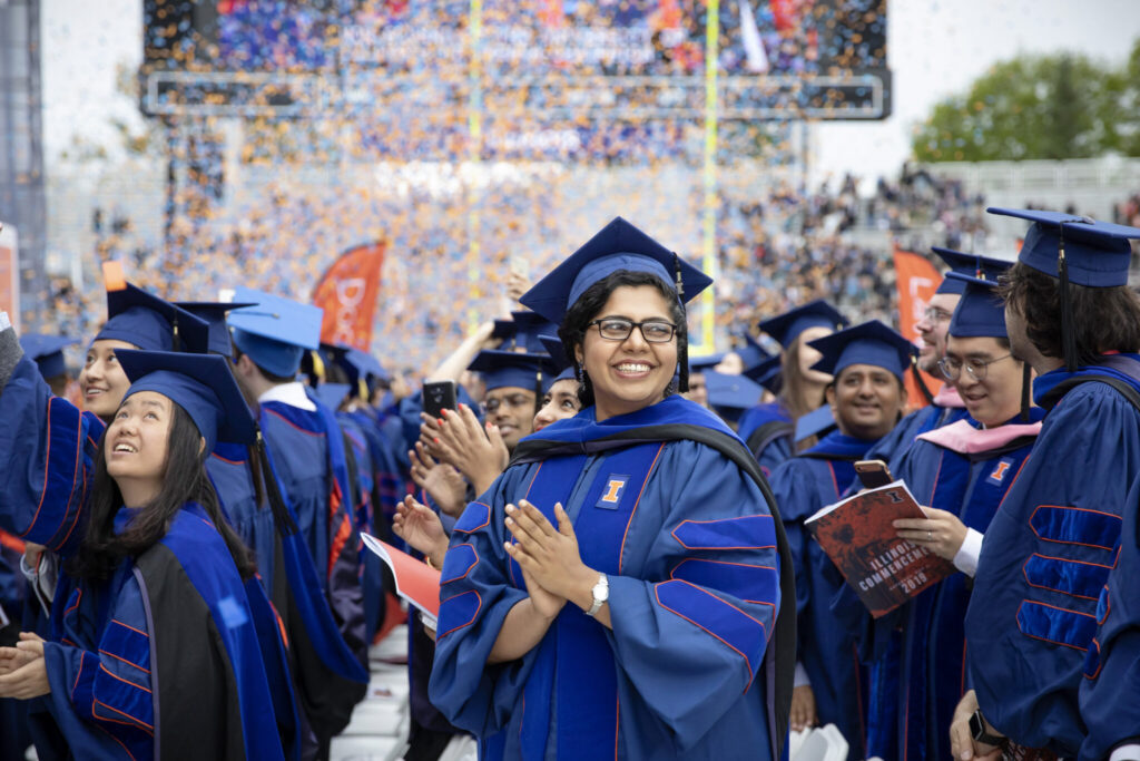 Dozens of University of Illinois graduates in blue academic regalia celebrating at their commencement ceremony, with confetti flying in the air. One student in the center of the frame clasps their hands together and smiles at fellow graduates. 