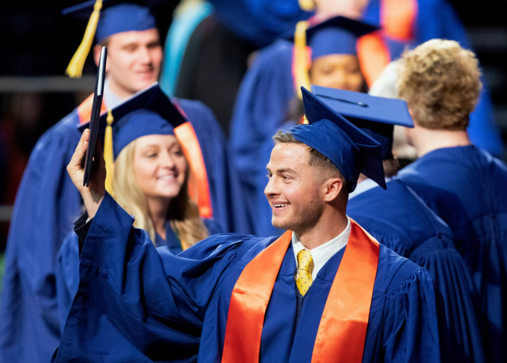 Graduates in regalia celebrating at a graduation ceremony, with one person in the foreground smiling and holding up a diploma.